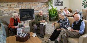 A group of four seniors sit and have coffee and chat in front of the fireplace