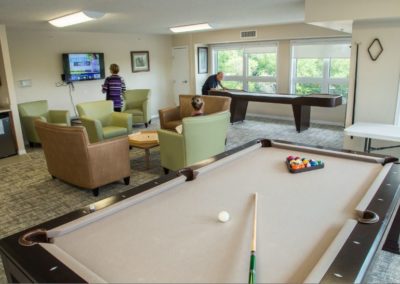 A man plays shuffleboard while a lady sits and reads a book while another lady changes the channel on the TV.